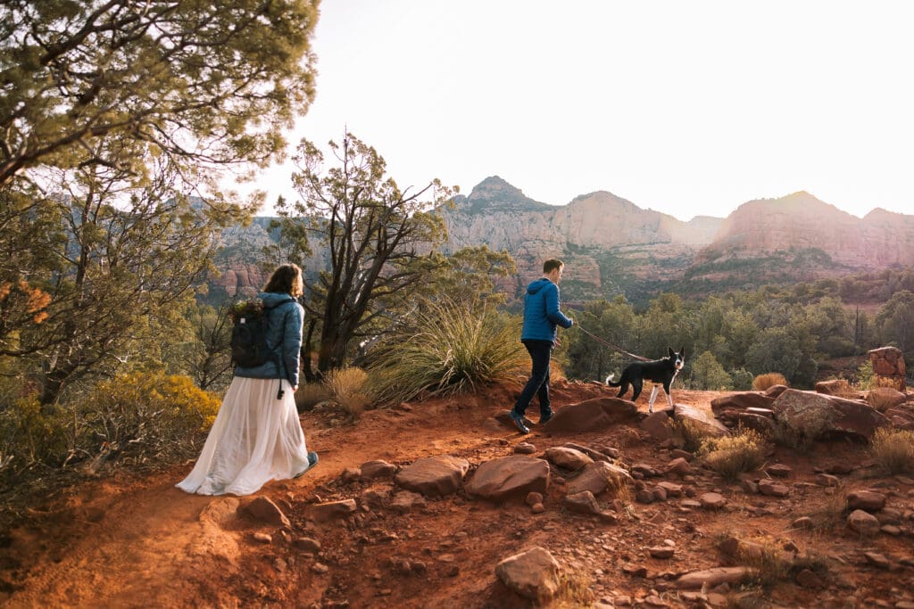 A bride and groom trudge up to their elopement location in jackets with their dog leading the way. 