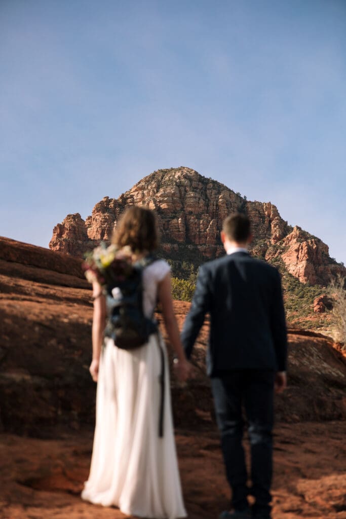 A bride and groom face a red rock, admiring it from a distance.
