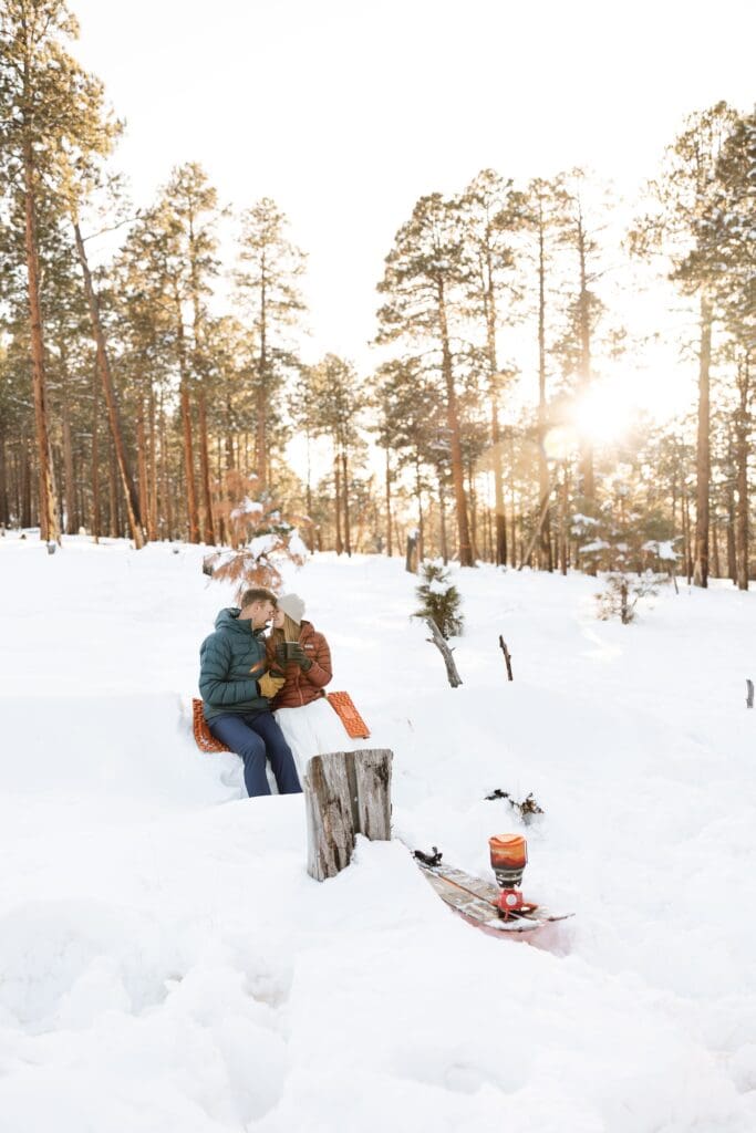 A bride and groom sit on a log as they hold each other and enjoy hot chocolate.