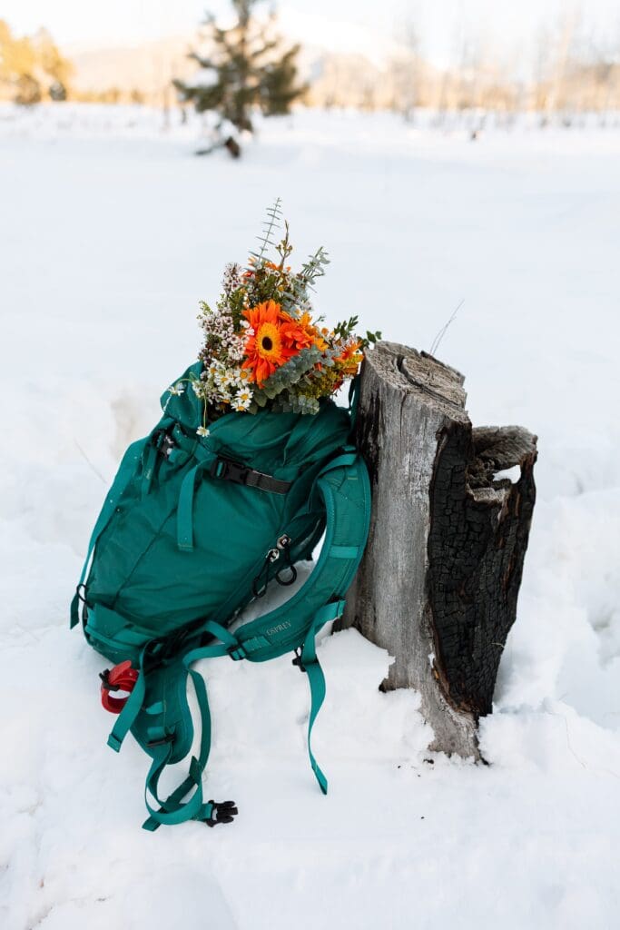 A brides bouquet is nestled in her day pack, leaning up against a snow covered log.