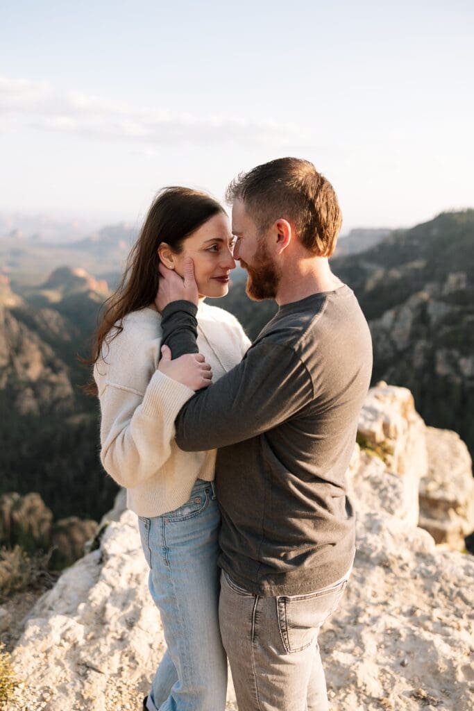 A couple stands facing each other while he holds her face gently in one hand with beautiful Sedona in the distance.