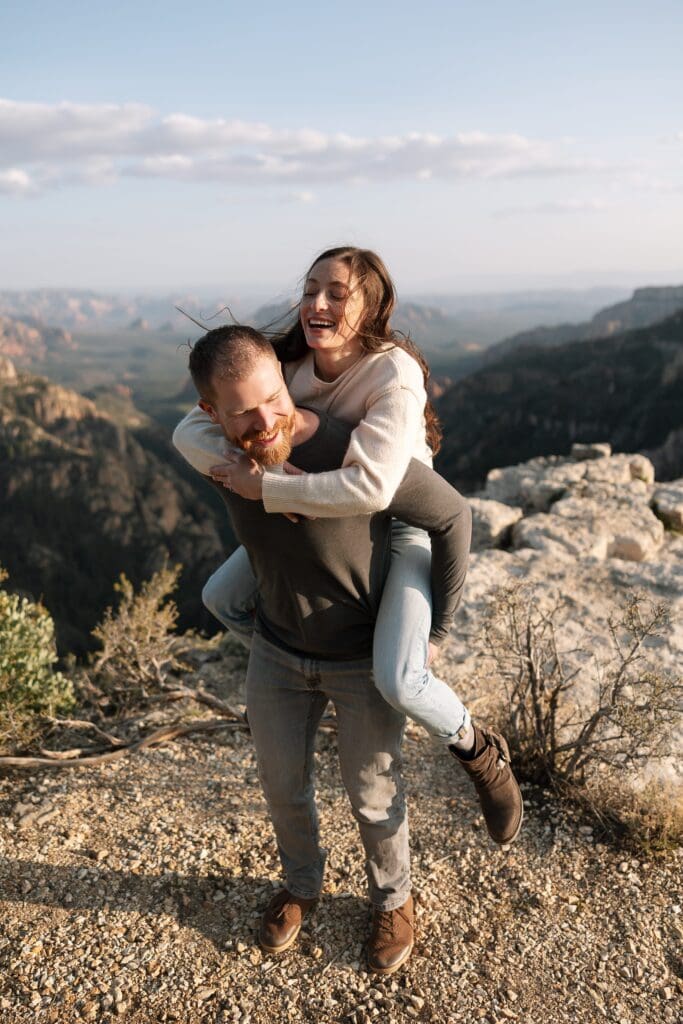 A man is giving his partner a piggie back ride with beautiful Sedona in the distance.