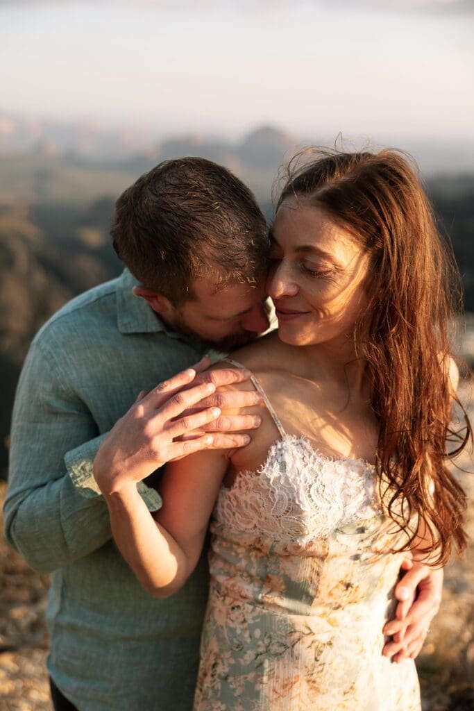 A man stands behind his partner kissing her neck softly with beautiful golden hour light on her face and body skin and Sedona blurred in the background.