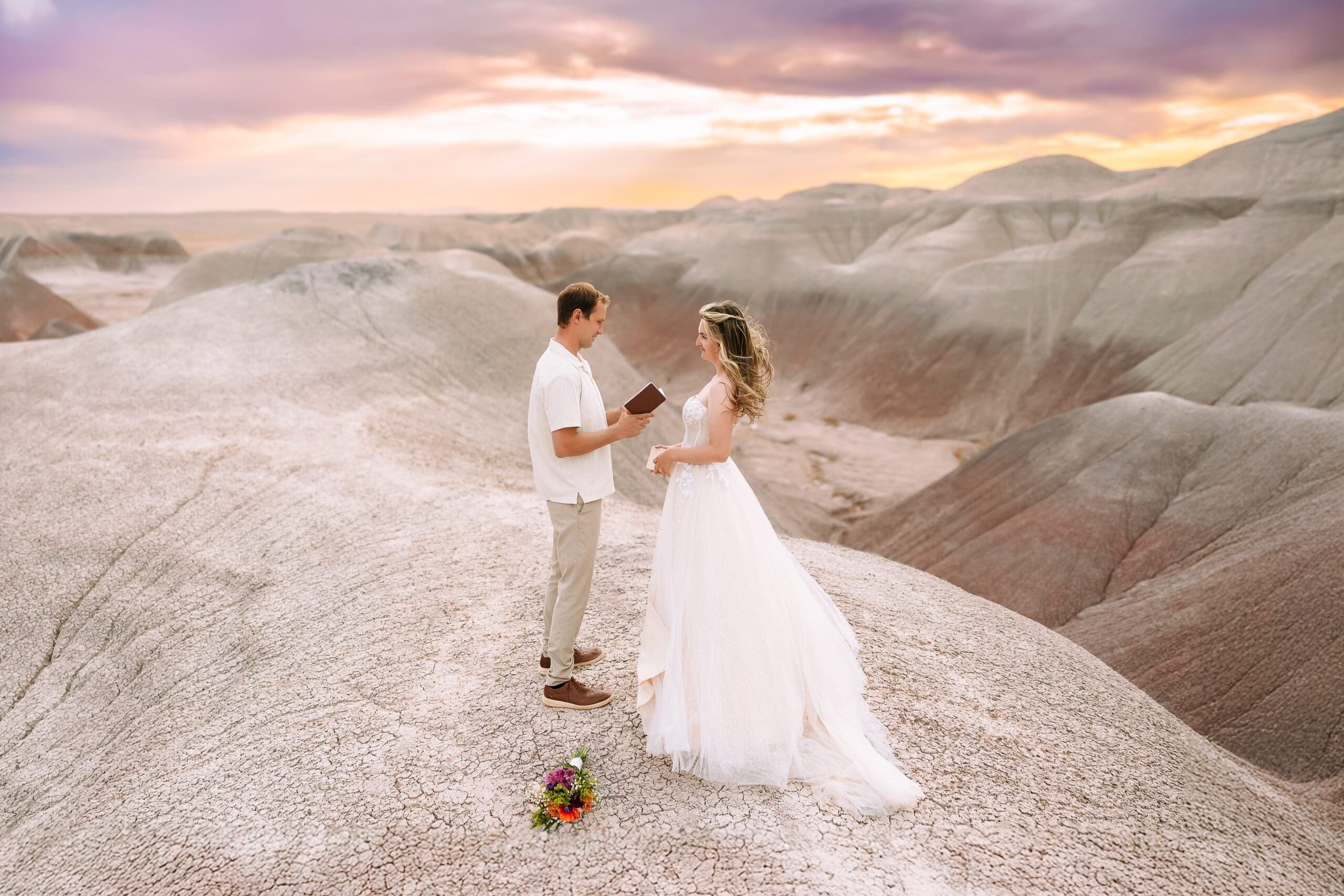 A bride and groom exchange vows while standing atop a desert mountain with pink and brown hues as the sun sets behind them.