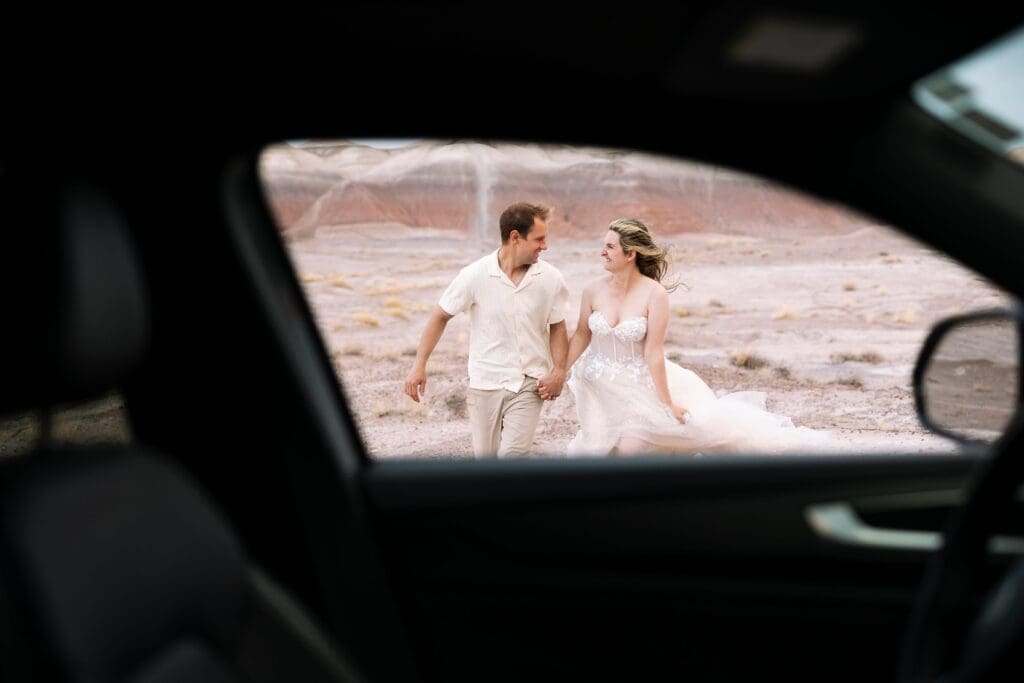 A bride and groom run toward the camera framed by the window of the car. 