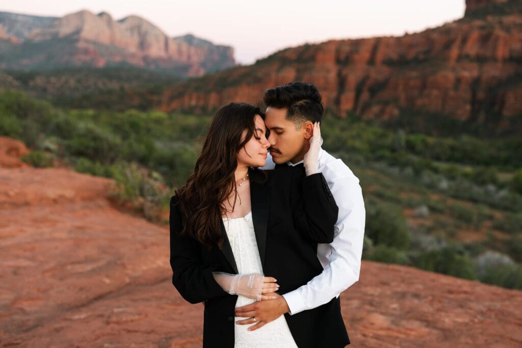 Bride wearing groom's jacket holding his face softly with Cathedral Rock and red rocks in the background during a Secret Slickrock Sedona elopement