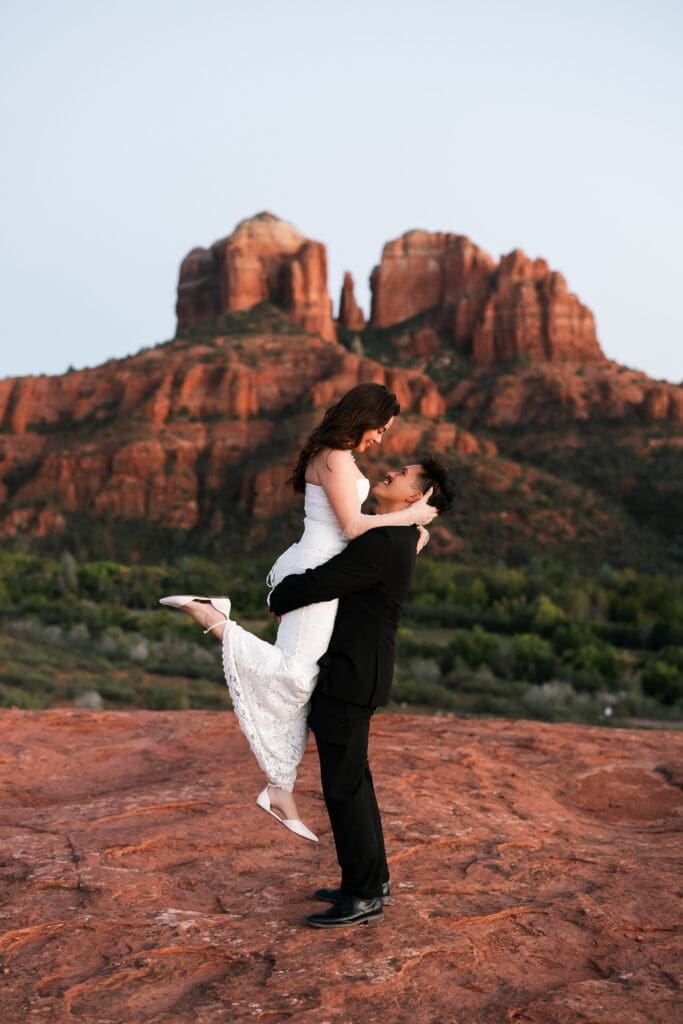 A groom lifts his bride with cathedral rock in the background at sunset.