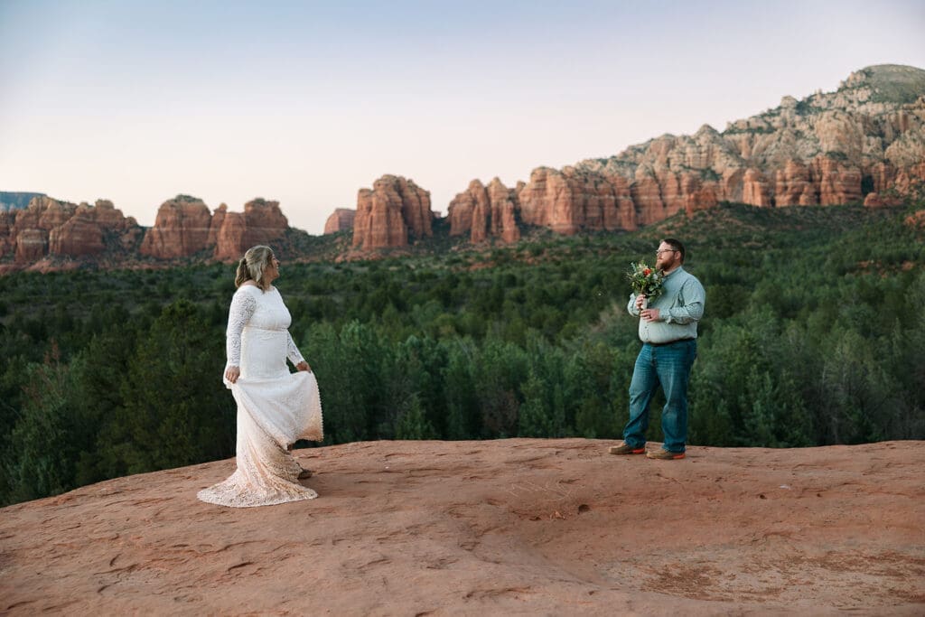Elopement couple standing on red rock cliffs overlooking Sedona canyons while the bride dances.