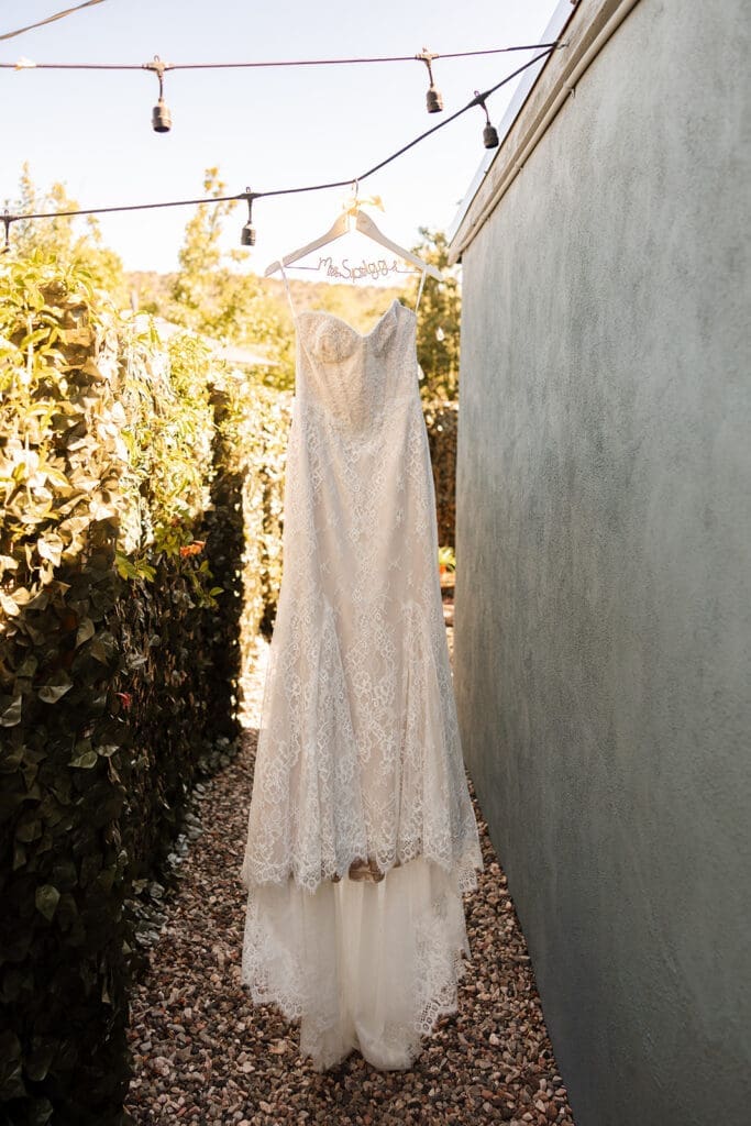 Bridal gown hanging on a wooden hanger against a neutral outdoor wall with overhead string lights at a Sedona rental home.