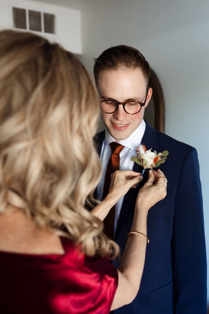 Intimate moment of the groom’s mother placing a floral boutonnierre on his navy blue suit jacket prior to the ceremony.
