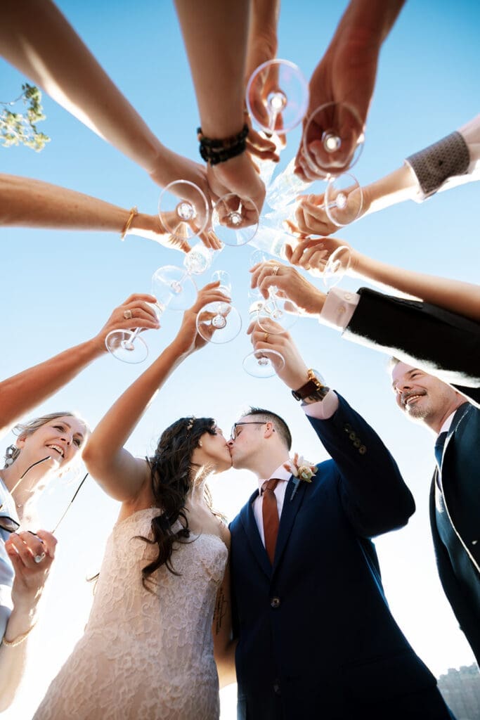 Bride and groom kissing while raising champagne glasses with family members in a low-angle celebratory group toast.