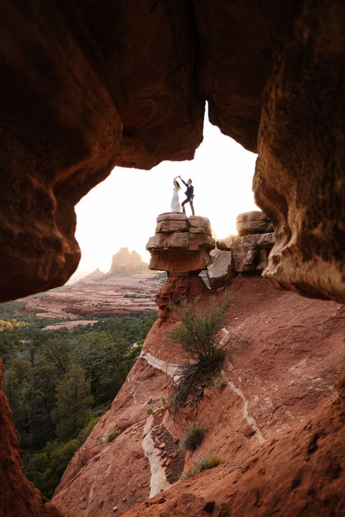 Wide-angle shot framing the bride and groom standing together inside the dramatic natural red rock cave opening in Sedona.