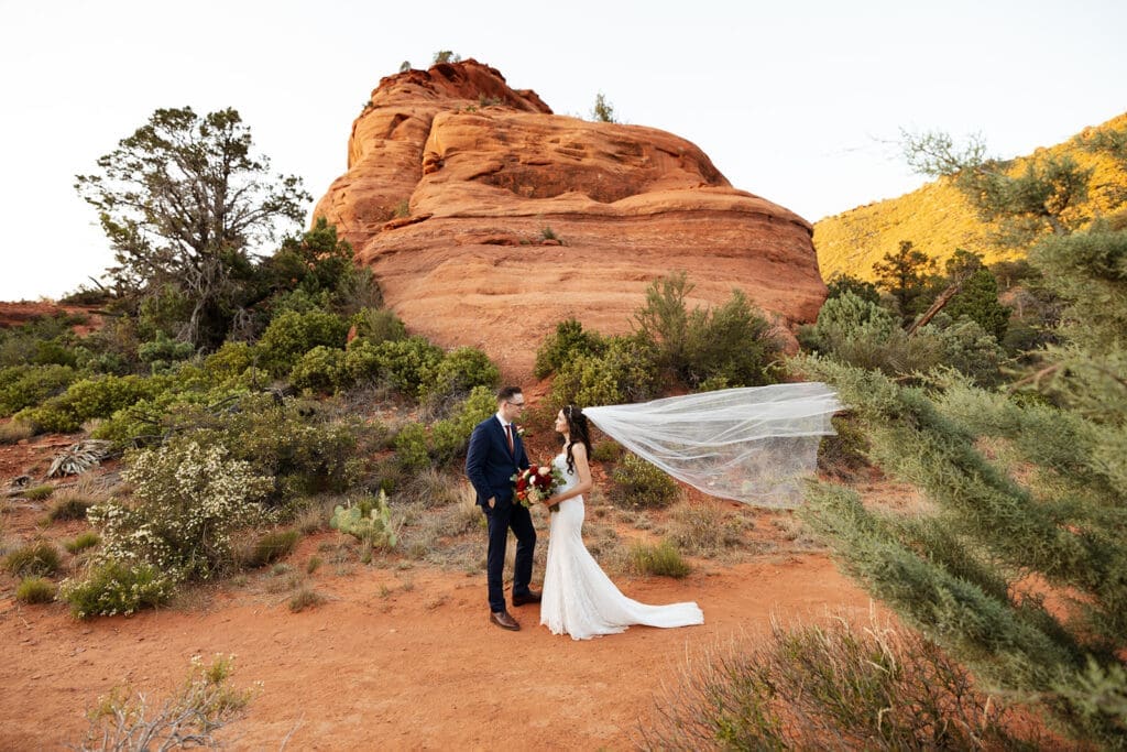 Wide-angle shot of the bride and groom standing together on a red rock cliffside with a sweeping desert mountain backdrop.