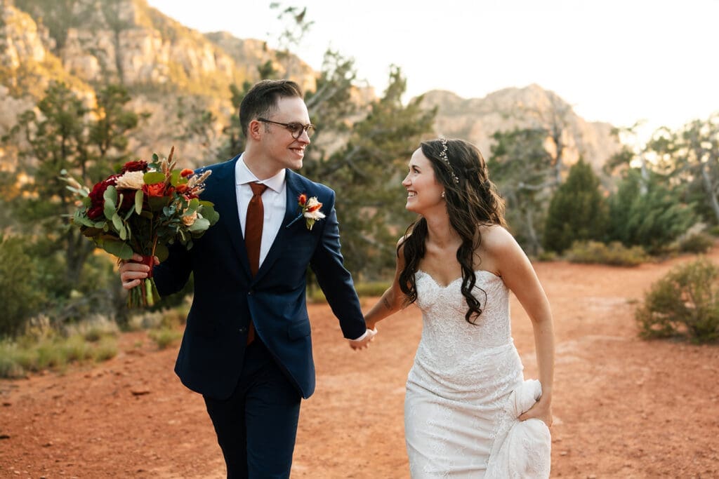 Smiling bride and groom holding hands and walking along a rugged trail during golden hour for their off-roading Sedona elopement.
