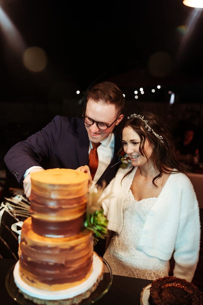 Eloping couple smiling together while slicing their multi-tiered rustic wedding cake decorated with desert greenery.