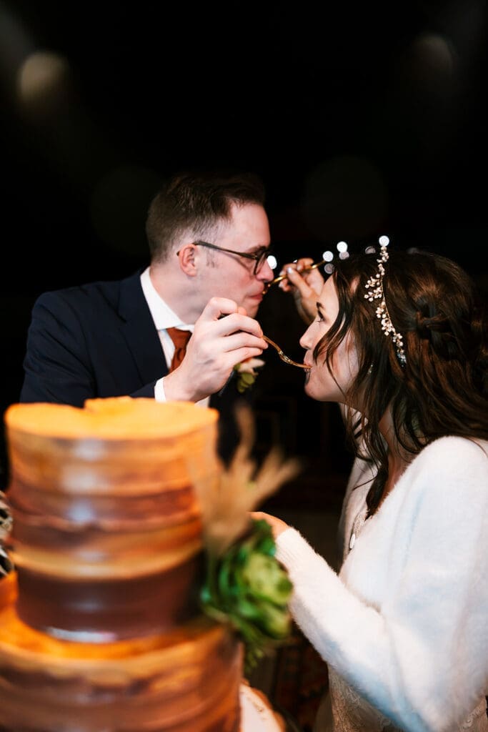 Groom with glasses playfully offering a bite of wedding cake to his bride at their intimate outdoor reception.