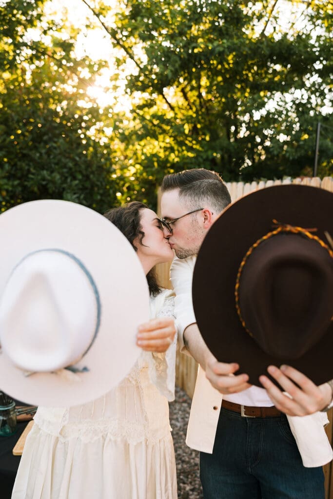 Candid shot showing a couple sharing an intimate moment and a kiss while showing off their newly customized hats at a pre-elopement party.