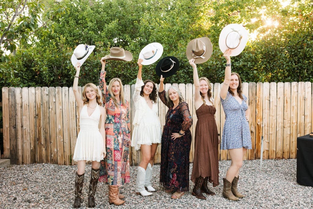 Happy bridesmaids laughing and holding up their customized cowboy hats outside against a rustic fence line before the Sedona elopement.