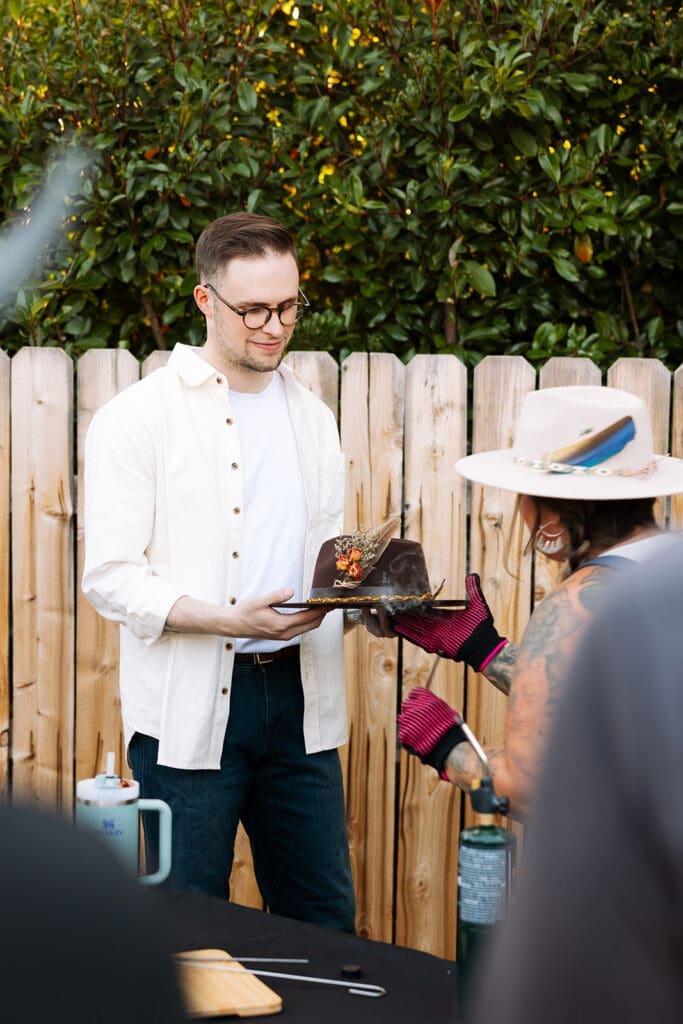 The groom is holding a personalized cowboy hat as the mobile hat bar owner, Rachel, brands his custom hat at an outdoor pre-elopement celebration.