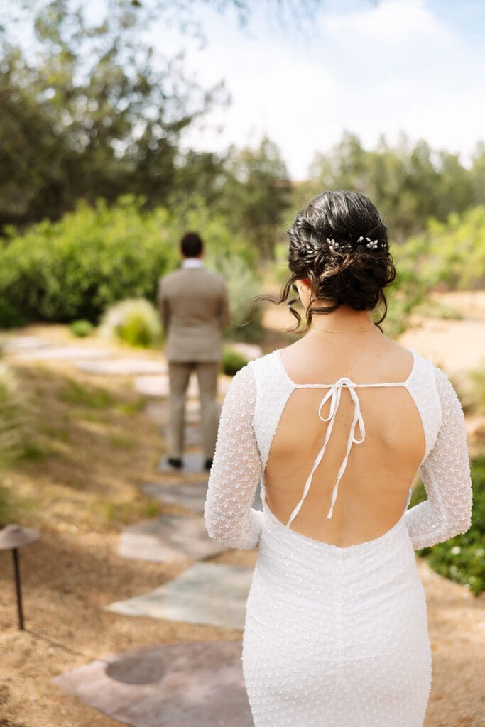 Back view of a bride in a long-sleeved lace wedding dress walking down a flagstone path toward the groom for a Sedona sunset elopement.