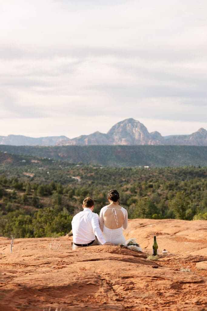 Bride and groom sitting together on a high red rock ledge overlooking the expansive Sedona valley and mountains sharing a picnic.