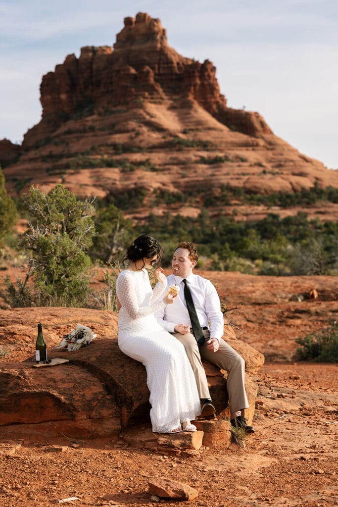 Newlyweds sitting side-by-side on red rocks sharing a champagne toast right after their Sedona ceremony.