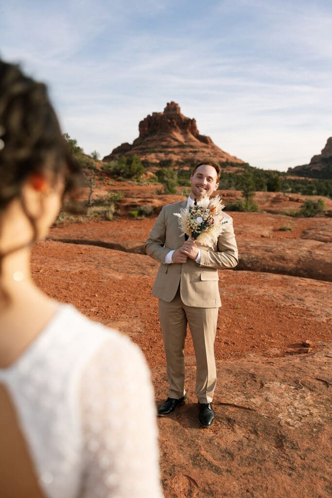Groom holding a bridal bouquet and smiling while looking over his shoulder at his bride in Sedona.