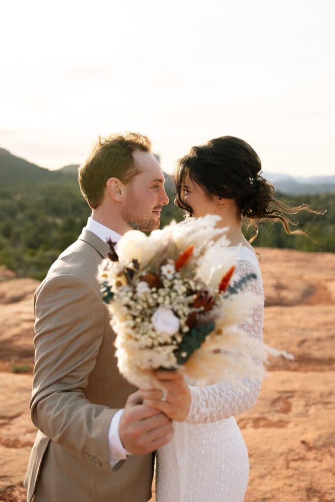 Smiling groom holding his bridal bouquet next to his bride against red rock formations during a Sedona sunset elopement.