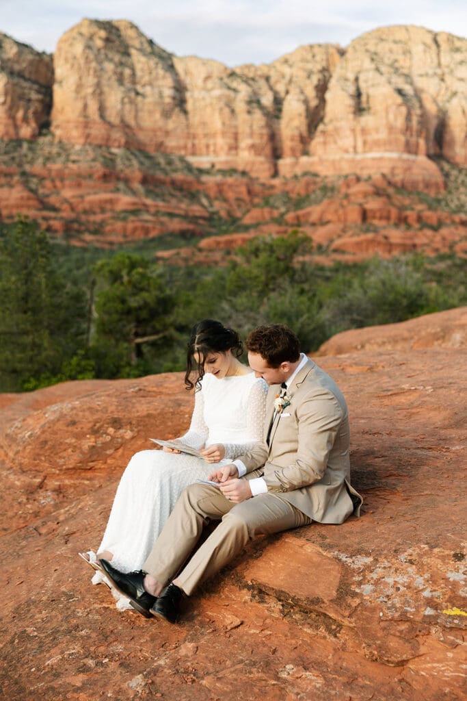 Bride and groom sitting side-by-side on the red dirt reading personalized letters to each other.