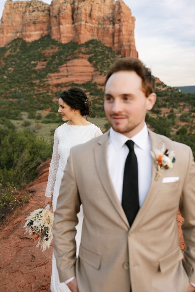 Vertical portrait of the groom in a tan suit with the bride in a long-sleeved lace gown standing closely behind him against a red rock backdrop.