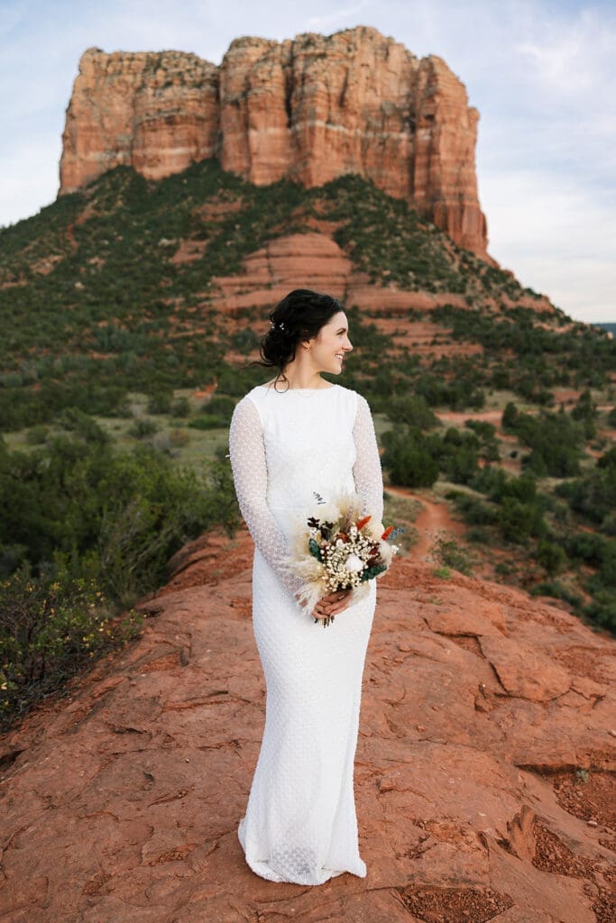 Full-length vertical portrait of the bride standing alone on a red rock ledge, holding her white and pampas grass bouquet.