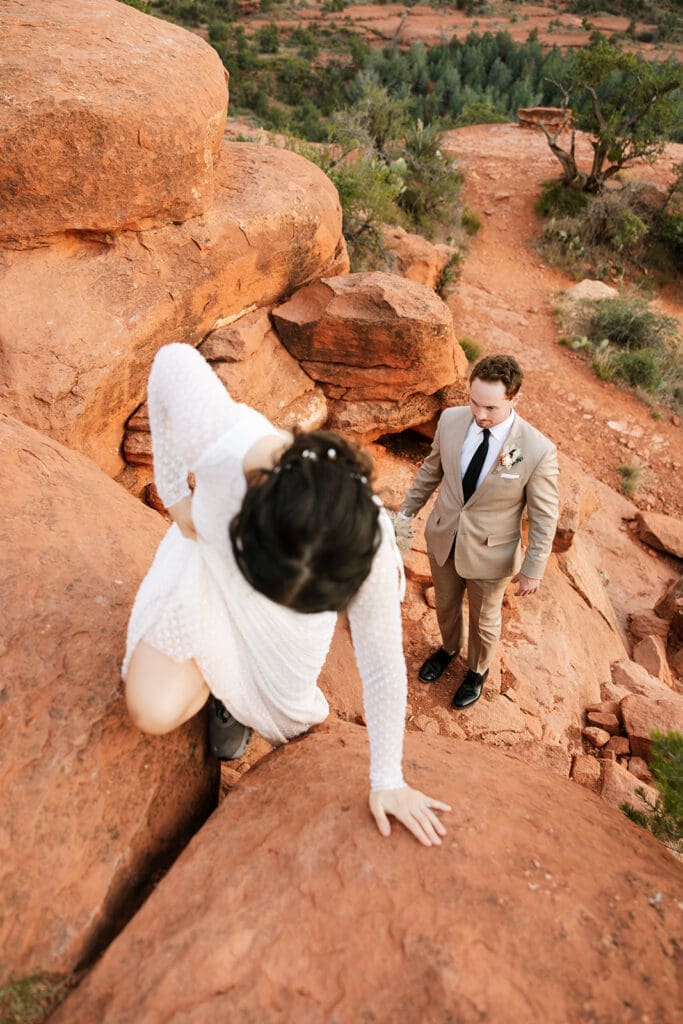 High-angle shot of the bride carefully climbing up a steep red rock cleft while the groom watches from below during their Sedona elopement.