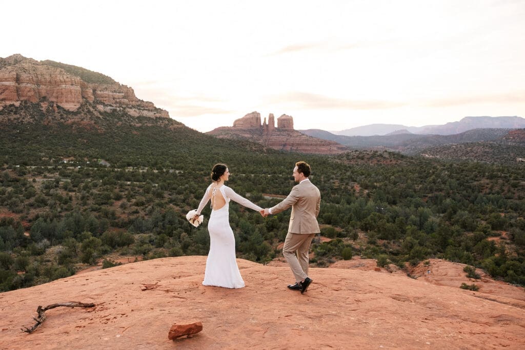 Wide horizontal landscape shot of the couple holding hands and walking across an expansive red rock plateau at sunset in Sedona.