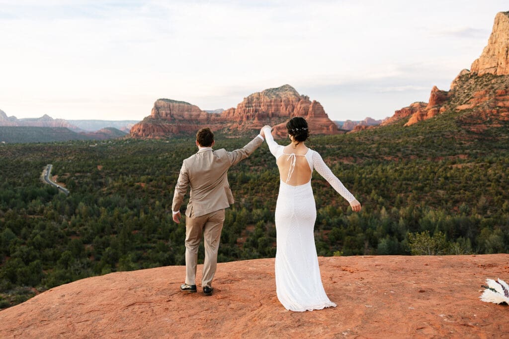Bride and groom holding hands and raising their arms playfully while walking along a red rock cliffside during a Sedona sunset elopement.