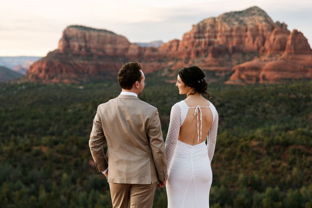 Back view of the groom in a tan suit standing next to his bride in a backless lace wedding gown, looking out at the Sedona landscape.