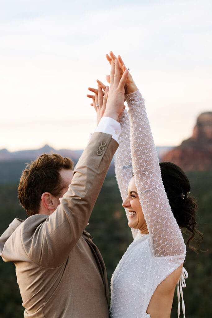Groom holding the bride's hand high as they share a joyful, laughing dance on the red rocks under a colorful twilight sky.