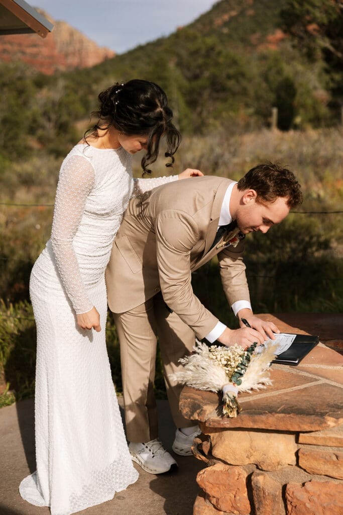 Bride and groom leaning over together to sign their marriage certificate on a rocky ledge during their Sedona sunset elopement.