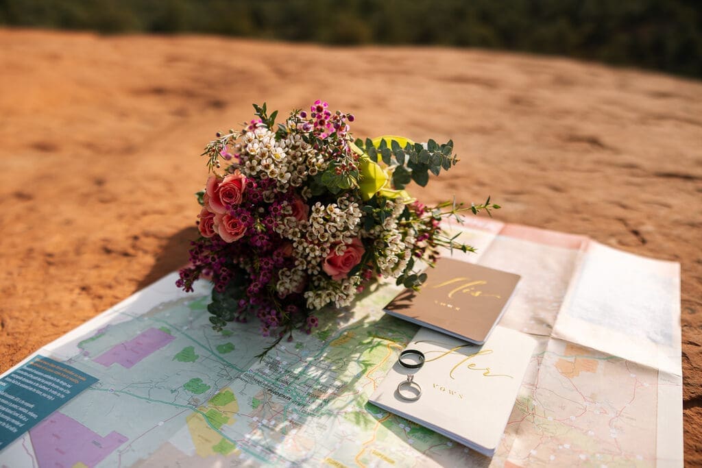 An elopement flat lay that includes a trail map, rings, vow books and florals.